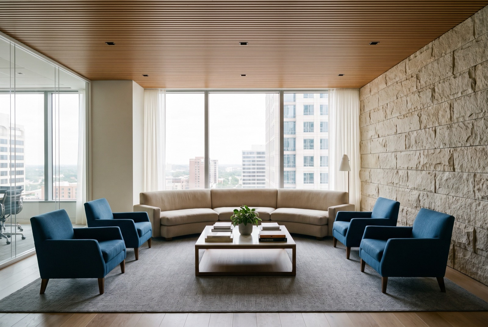 A lounge with blue armchairs, a stone wall and a city view through full-height glass.